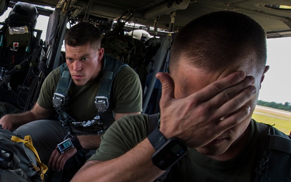 Military personnel, appearing to be U.S. Marines based on visible MARPAT camouflage uniforms and gear, are seated inside a military helicopter. The foreground figure has his hand covering his face, while another service member in the background looks directly at the camera. The aircraft appears to be on or near a runway or airfield, visible through the open door.