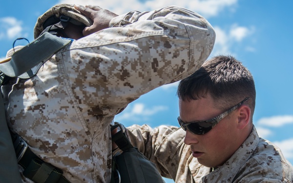 Two U.S. Marines in MARPAT desert camouflage uniforms are on a tarmac or airfield, with one Marine inspecting or adjusting a parachute rig on the other. The Marine performing the check has a name tape partially visible reading 'SSETT' (likely 'BASSETT' or similar) and the word 'MARINE' on his uniform, and is wearing sunglasses. A third Marine is visible in the background walking away.