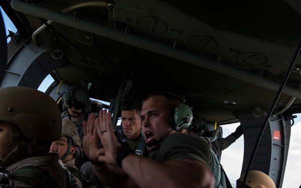 Military personnel are seated inside a military helicopter or transport aircraft, appearing to be preparing for a parachute jump operation. A crew member wearing a flight helmet and headset is giving hand signals or instructions to the seated soldiers, who are wearing combat helmets and parachute harnesses. The open side of the aircraft reveals an aerial view of terrain and clouds below.