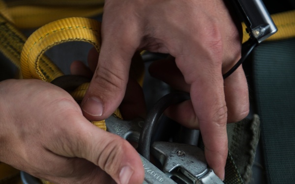 A person is handling safety or rigging hardware, specifically connecting or inspecting metal snap hooks and carabiners attached to webbing straps. The image shows close-up detail of hands manipulating load-bearing connectors on what appears to be a harness or tie-down system. Yellow and green nylon webbing straps are visible alongside the metal hardware.