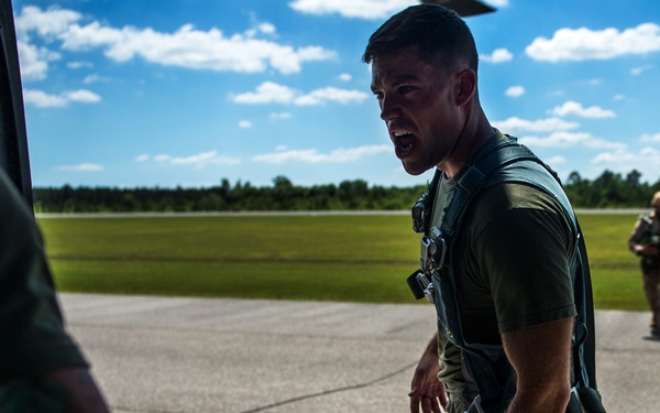 A military personnel member wearing a tactical harness/vest is shouting or giving instructions near what appears to be the rear ramp of a military aircraft on a tarmac. At least two other personnel are visible in the background, also in military attire. The setting appears to be a military airfield during daytime with clear skies.