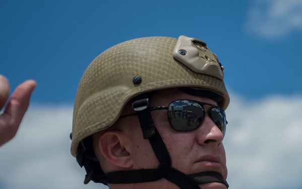 A military service member is photographed in close-up against a blue sky with scattered clouds. The individual is wearing standard combat gear including a tan/desert-colored ballistic helmet and MARPAT desert camouflage uniform. Both hands are raised, suggesting the person may be giving hand signals or instructions.