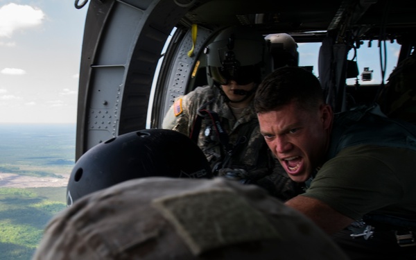 Military personnel are inside an open helicopter at altitude, with a forested landscape visible below. One individual in a green shirt appears to be shouting commands or instructions, while others in military uniforms and helmets are positioned near the open door. The scene suggests a airborne training or jump operation in progress.