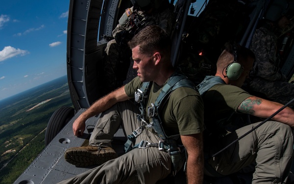 Two military personnel in olive drab uniforms are seated on the open ramp of a military helicopter in flight, with a forested landscape visible far below. Both individuals are wearing safety harnesses and appear to be conducting an aerial operation or training exercise. A green equipment bag with yellow straps is visible on the ramp between them.
