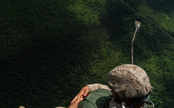 A military service member in USMC desert MARPAT camouflage uniform is exiting or has just exited an aircraft, visible at the lower left edge of the frame. The individual is in a tucked body position with a static line visible trailing upward, indicating a static-line parachute jump. Dense green forested terrain is visible far below, suggesting significant altitude.