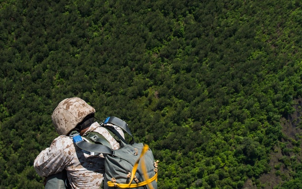 A military service member in desert camouflage (MARPAT) uniform is positioned at the edge of an aircraft, preparing to conduct a parachute jump over a densely forested, mountainous terrain. The individual is equipped with a parachute pack and is connected to a yellow static line attached to the aircraft. The lush green landscape below suggests a tropical or subtropical environment.