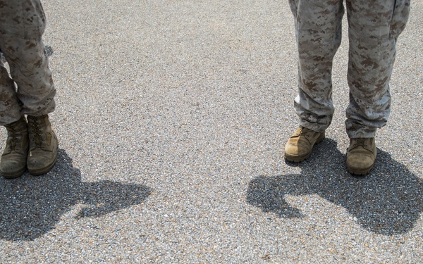 The image shows the lower legs and boots of two individuals wearing military camouflage uniforms, standing on a gravel or asphalt surface. Their shadows are cast prominently on the ground, suggesting bright overhead sunlight. The shadows reveal the individuals are holding rifles or similar long weapons at their sides.