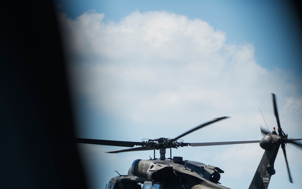 A military utility helicopter, consistent with a UH-60 Black Hawk, is photographed in flight through what appears to be a window or opening frame. The helicopter is dark/olive drab in color and has at least one crew member or soldier visible hanging or standing at the open side door. The image is captured from inside another aircraft or structure, giving a framed perspective against a partly cloudy blue sky.