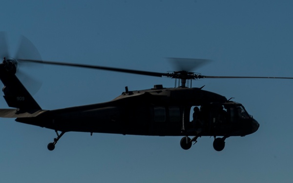 A military helicopter is captured in silhouette against a clear blue sky with scattered clouds near the horizon. The aircraft appears to be a UH-60 Black Hawk type helicopter in flight, photographed from a low angle. The strong backlighting creates a dramatic dark silhouette effect, obscuring most surface details.