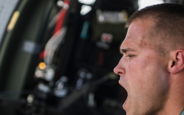 A young military personnel member is seated inside what appears to be a military aircraft or helicopter, with his mouth open as if shouting or communicating. He is wearing a green military flight suit and a tactical vest, and appears to be holding a yellow strap or tether. The background shows the interior cockpit or cargo area of the aircraft with various equipment and controls visible.
