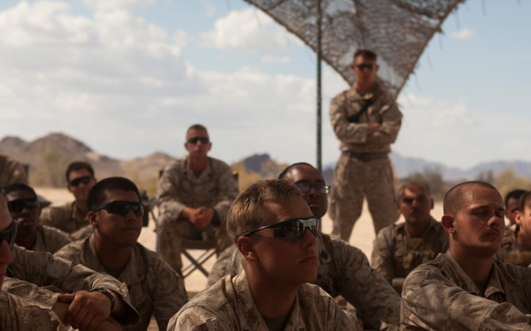 A group of U.S. Marines in desert camouflage (MARPAT) uniforms are seated on the ground in an arid, desert environment, appearing to receive a briefing or instruction. One Marine stands in the background with arms crossed beneath a camouflage netting structure. The setting suggests a field training exercise or operational briefing in a desert location.
