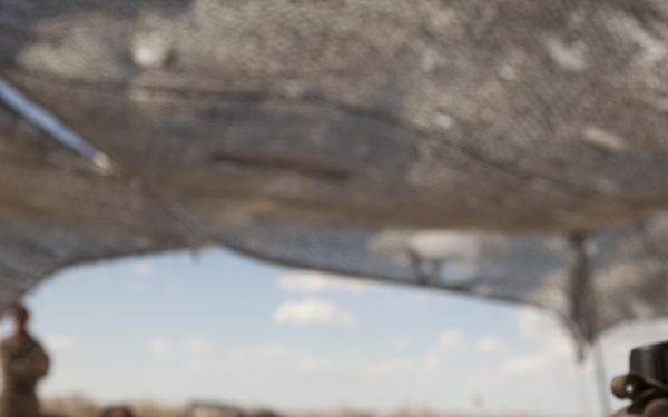Military personnel are gathered under a camouflage netting shade structure in an arid, desert-like environment. In the foreground, numerous small plastic cups filled with a dark red liquid are arranged on a table alongside a military-issue canteen. A service member in camouflage uniform stands at the front, appearing to address the seated group.