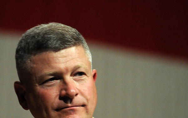 A U.S. Navy officer in khaki service uniform is photographed against what appears to be an American flag backdrop. His name tag reads 'RICK WEST' and he displays multiple insignia, badges, and service ribbons on his uniform. He appears to be at an official military event or ceremony.