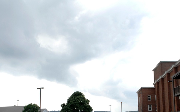 A large formation of military personnel in Navy Working Uniform (NWU) blue digital camouflage are marching across a crosswalk on what appears to be a military installation. Several observers in different uniforms, including woodland camouflage and khaki, stand to the right watching the formation. The scene takes place under overcast, stormy skies near brick institutional buildings.