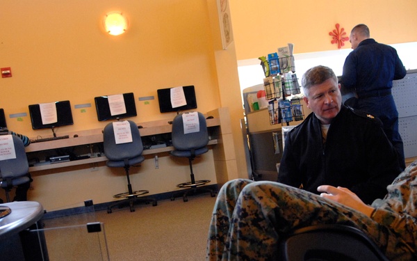 Two individuals are seated in an office or computer lab environment, engaged in conversation. One person is wearing United States Marine Corps MARPAT camouflage uniform with tan combat boots, while the other is in civilian dark clothing. A row of desktop computers with office chairs is visible in the background, along with a third person standing at a counter.