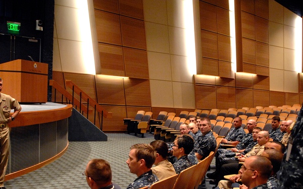 A military briefing or meeting is taking place in a modern auditorium. A senior officer in tan/khaki Navy service uniform stands at the front near a podium, addressing a group of seated personnel predominantly wearing Navy Working Uniform (NWU) digital camouflage. A second uniformed individual stands at the right side of the frame observing the audience.