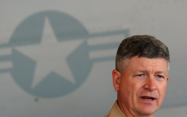 A U.S. Navy officer identified by his name tag as 'Rick West' is speaking at a microphone, appearing to deliver a speech or address. He is wearing a tan/khaki Navy service uniform with various insignia, ribbons, and badges. Behind him is a military aircraft bearing a U.S. military roundel (star-and-bar insignia).