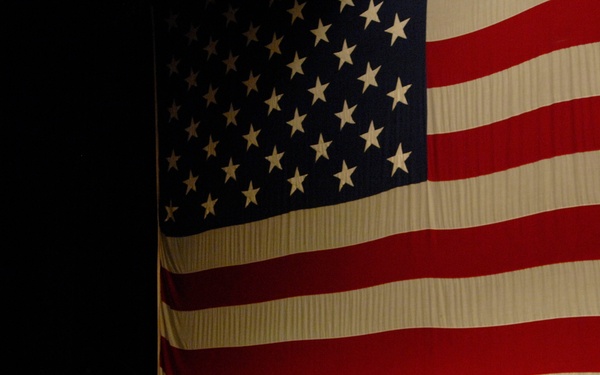 A U.S. Navy officer in khaki service uniform stands on a stage speaking or presenting, with a large American flag displayed as a backdrop. The setting appears to be an auditorium or theater with a wooden stage floor. The officer appears to be gesturing with his hands, suggesting an address or briefing to an audience.