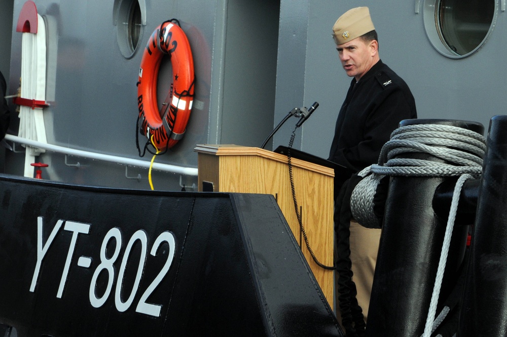 A naval officer in dress uniform stands at a wooden podium speaking at what appears to be a military commissioning or decommissioning ceremony aboard a vessel. The hull marking 'YT-802' is prominently visible, identifying this as a U.S. Navy yard tug. An orange life ring is mounted on the gray ship's superstructure in the background.