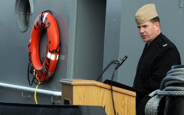 A naval officer in dress uniform stands at a wooden podium speaking at what appears to be a military commissioning or decommissioning ceremony aboard a vessel. The hull marking 'YT-802' is prominently visible, identifying this as a U.S. Navy yard tug. An orange life ring is mounted on the gray ship's superstructure in the background.