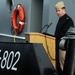 A naval officer in dress uniform stands at a wooden podium speaking at what appears to be a military commissioning or decommissioning ceremony aboard a vessel. The hull marking 'YT-802' is prominently visible, identifying this as a U.S. Navy yard tug. An orange life ring is mounted on the gray ship's superstructure in the background.