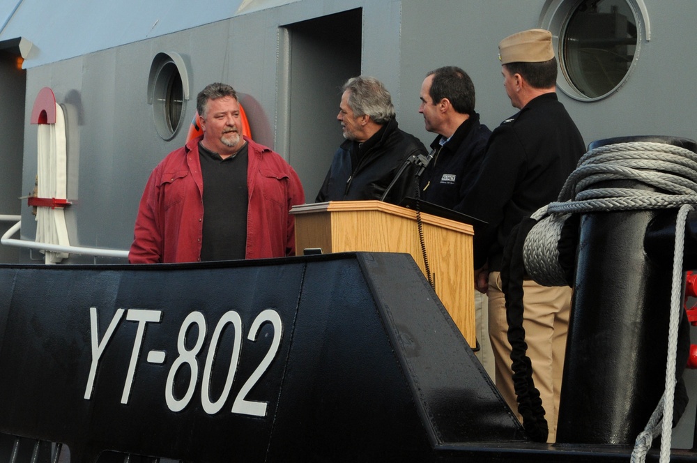 A group of four men are gathered on or near a naval vessel designated YT-802, a yard tug classification. Three men stand near a wooden podium/lectern, suggesting an official ceremony or commissioning event, while one man in a red jacket stands slightly apart. A uniformed military personnel wearing a garrison cap is present among the group.