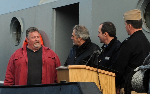 A group of four men are gathered on or near a naval vessel designated YT-802, a yard tug classification. Three men stand near a wooden podium/lectern, suggesting an official ceremony or commissioning event, while one man in a red jacket stands slightly apart. A uniformed military personnel wearing a garrison cap is present among the group.