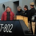 A group of four men are gathered on or near a naval vessel designated YT-802, a yard tug classification. Three men stand near a wooden podium/lectern, suggesting an official ceremony or commissioning event, while one man in a red jacket stands slightly apart. A uniformed military personnel wearing a garrison cap is present among the group.