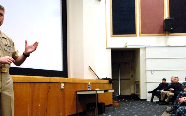 A senior U.S. Navy officer in tan Service Khaki uniform is addressing a group of sailors seated in an auditorium or briefing room. The audience members are wearing Navy Working Uniform (NWU) blue digital camouflage pattern. The setting appears to be a formal military briefing or all-hands meeting at a naval installation.