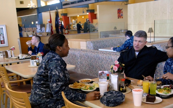 Three individuals are seated at a table in what appears to be a military dining facility (mess hall or cafeteria). Two individuals are wearing U.S. Navy blue digital camouflage uniforms (NWUs), while a third person in a dark civilian or officer jacket is engaged in conversation with them. Food trays, drinks, and plates of food are visible on the table.
