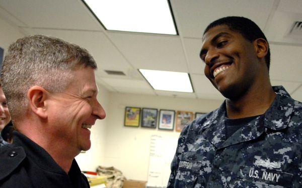 Three individuals are gathered in what appears to be a U.S. Navy administrative or office space. Two are wearing U.S. Navy NWU (Navy Working Uniform) digital camouflage uniforms, while one person in the foreground wears a dark service uniform. All three appear to be engaged in a positive, lighthearted interaction.