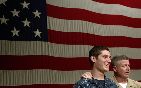 Two U.S. Navy personnel stand in front of a large American flag backdrop. The younger sailor in digital blue camouflage (NWU Type I) with the name tape 'VITALE' is being recognized or acknowledged by a senior officer in khaki service uniform who has his arm around the sailor's shoulder. The setting appears to be a formal military ceremony or event.