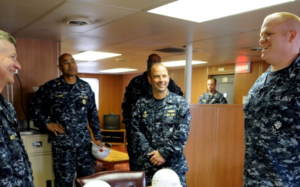 A group of U.S. Navy personnel in Navy Working Uniform (NWU) digital camouflage are gathered in what appears to be a shipboard conference or briefing room. Two individuals in the foreground appear to be engaged in a conversation or informal exchange, both smiling. Two white hard hats and a pair of safety glasses are placed on the wooden conference table, suggesting a safety or inspection-related context.