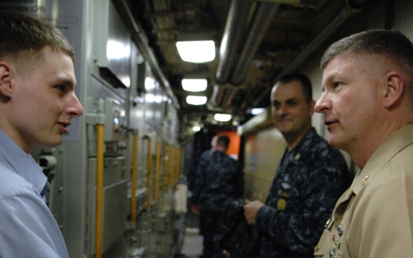 A young man in a light blue dress shirt is shaking hands with a senior military officer in a tan/khaki uniform with visible ribbons and insignia. The setting appears to be the interior corridor of a naval vessel, with other personnel in Navy working uniforms visible in the background. The interaction suggests a formal recognition or greeting ceremony.