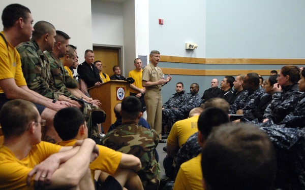 A military briefing or all-hands meeting is taking place in a large room, with a senior naval officer in dress uniform speaking at a podium. The audience consists of a mixed group of personnel wearing Navy Working Uniform (NWU blue digital camouflage), yellow physical training shirts, and woodland camouflage uniforms. The room is packed, with some attendees seated on the floor due to limited seating.