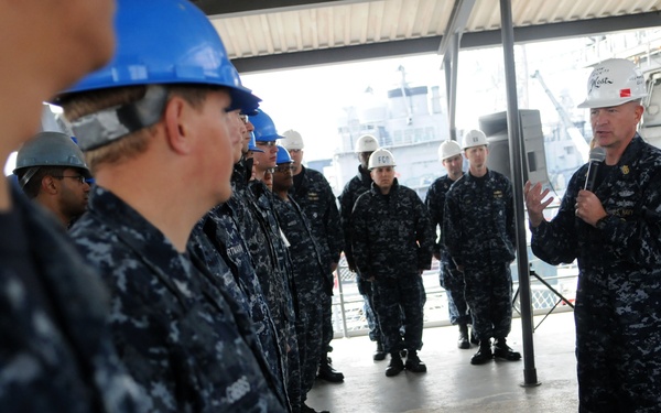 A senior U.S. Navy officer wearing a white hard hat is addressing a group of sailors in Navy Working Uniform (NWU) Type I (blue digital camouflage) who are wearing blue and white hard hats. The setting appears to be a covered pier or shipyard facility, with a naval vessel visible in the background. The scene suggests a formal briefing or all-hands meeting in a shipyard or maintenance environment.