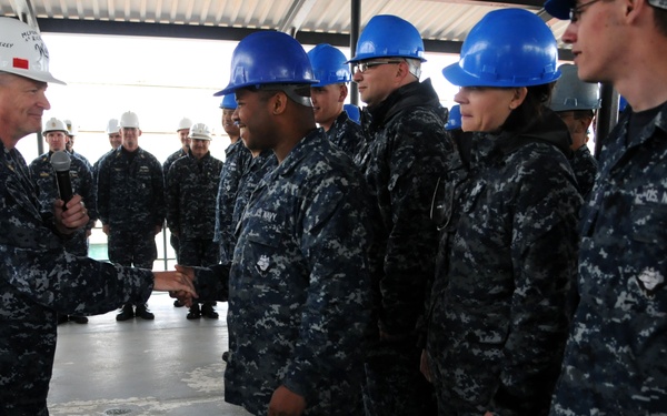 A senior U.S. Navy officer wearing a white hard hat is shaking hands with personnel wearing blue hard hats and Navy Working Uniforms (NWU). The setting appears to be an industrial or shipyard facility, likely aboard a vessel under maintenance or construction. The scene suggests an official visit or inspection by a high-ranking officer.