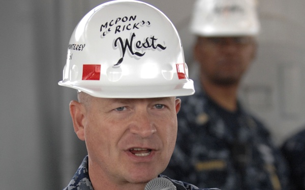 A U.S. Navy enlisted sailor in Navy Working Uniform (NWU) digital camouflage is speaking into a Shure microphone while wearing a white hard hat inscribed with 'MCPON Rick West' and partially visible text 'MONTEREY'. The individual appears to be addressing an audience at an official Navy event or facility visit. A second person in Navy uniform is visible in the blurred background.