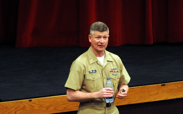 A senior U.S. Navy officer in khaki service uniform is addressing a seated audience of military personnel in what appears to be an auditorium or theater setting. The speaker is standing in front of a stage with a red curtain backdrop, holding a water bottle while speaking. The audience, visible from behind, consists of multiple uniformed service members.