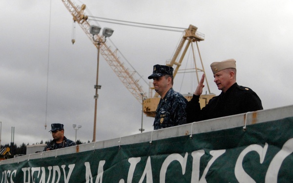 The image shows U.S. Navy personnel aboard or near the USS Henry M. Jackson (SSBN 730), a ballistic missile submarine, at what appears to be a naval base. A large green banner reading 'USS HENRY M JACKSON SSBN 730 DEFENDER OF FREEDOM' is prominently displayed on a gangway or brow structure. Several sailors in uniform are visible, with a large yellow crane visible in the background.