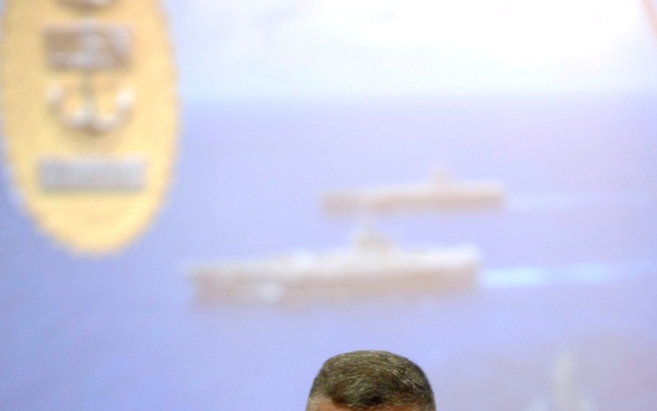 A U.S. Navy officer in Navy Working Uniform (NWU) Type I digital camouflage is delivering a presentation or briefing. Behind him is a projected slide featuring naval vessels and yellow text, along with what appears to be a Navy emblem or seal. The officer appears to be mid-speech, gesturing with his hands.