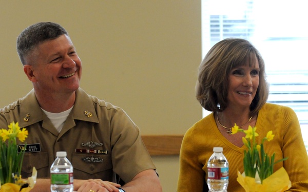 A U.S. Navy officer in khaki uniform and a woman in a yellow sweater are seated together at a white-clothed table, both smiling during what appears to be a formal or semi-formal gathering. The officer's name tag reads 'RICK WEST' and he displays several ribbons and a warfare qualification badge. The setting suggests a community or military family event.