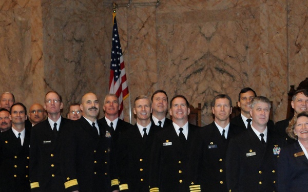 A group of approximately 20 individuals, predominantly wearing dark Navy military dress uniforms with gold buttons and rank insignia, pose for a formal group photograph in what appears to be a legislative chamber. Two civilian women in dark and white jackets stand at the right side of the group. An American flag is visible in the background, and legislative desks with nameplates are visible in the foreground.