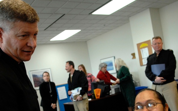 The image shows a military office setting where a senior officer in dark Navy uniform is interacting with a seated female sailor in tan/khaki Navy uniform. The seated sailor's name tag reads 'BASILIQ WALLY' and she is wearing service ribbons. Several civilians and military personnel are visible in the background engaged in various activities.