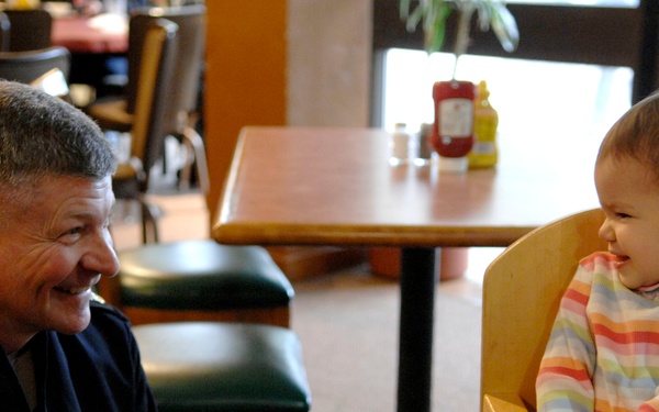 An adult male and a young infant are seated across from each other at a table in a casual restaurant setting. The man is smiling warmly at the baby, who is seated in a wooden high chair and appears to be laughing or smiling back. The interaction suggests a joyful, affectionate moment between the two.