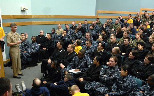 A large gathering of U.S. Navy personnel in an auditorium or briefing room, listening to a senior officer in dress uniform speaking at a podium. The audience includes sailors in various uniforms including Navy Working Uniform (NWU digital camouflage), physical training gear (yellow shirts), and camouflage utilities. The room is filled to capacity with personnel seated in chairs and on the floor.