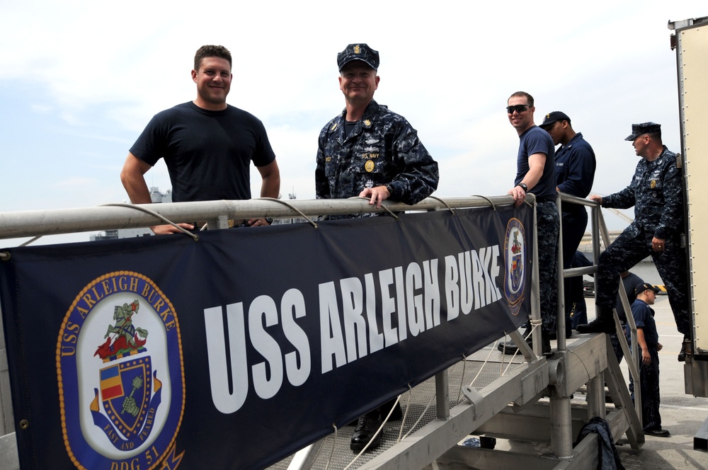 Several individuals, including uniformed U.S. Navy personnel and one civilian in a black t-shirt, are posing on a gangway or brow of a naval vessel. A prominent banner reading 'USS Arleigh Burke' with the ship's crest (DDG-51) is displayed on the railing. The setting appears to be a port or pier during daytime.