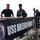 Several individuals, including uniformed U.S. Navy personnel and one civilian in a black t-shirt, are posing on a gangway or brow of a naval vessel. A prominent banner reading 'USS Arleigh Burke' with the ship's crest (DDG-51) is displayed on the railing. The setting appears to be a port or pier during daytime.