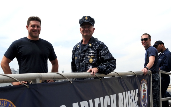 Several individuals, including uniformed U.S. Navy personnel and one civilian in a black t-shirt, are posing on a gangway or brow of a naval vessel. A prominent banner reading 'USS Arleigh Burke' with the ship's crest (DDG-51) is displayed on the railing. The setting appears to be a port or pier during daytime.