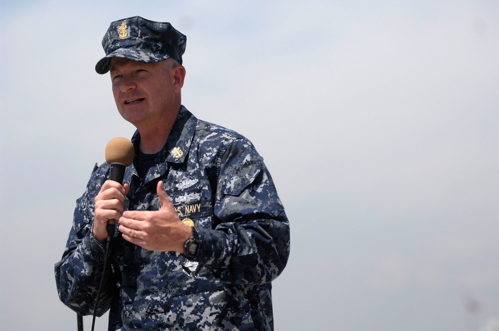 A U.S. Navy servicemember in Navy Working Uniform (NWU Type I) digital camouflage is speaking into a handheld microphone, likely addressing an audience at an outdoor event. The individual wears visible rank/rating insignia on their cap and chest, along with a qualification badge consistent with submarine warfare. The background is overcast and hazy, suggesting an outdoor setting near water or an open area.